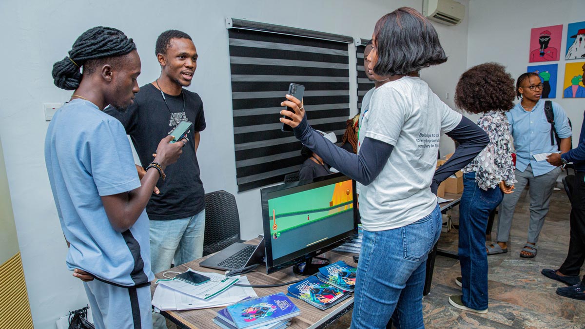 A group of people interacting in a tech event around a computer monitor showing a game.
